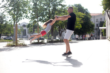 Father and daughter take fun outdoor in the street . Man throws up the girl to sky. The kid laughing and imagines what is flying.