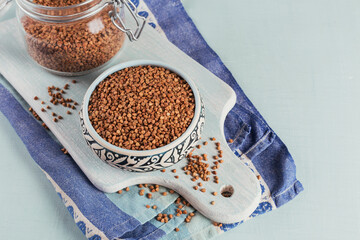 Bowl of dry raw buckwheat groats and glass jar on a light blue background. Cooking buckwheat porridge concept.