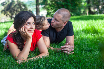 happy parents with daughter lies on the green grass.  People having fun on the park. Woman and kid hug the father