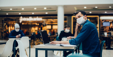 man in a protective mask works on a laptop sitting at a table in a cafe.