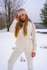 Winter lifestyle, young blonde caucasian girl with white winter outfit and woolen hat in the snow next to snowy pine trees, holidays in nature