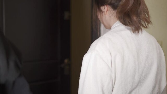 A young woman in a dressing gown opens the front door of the apartment. Back view.