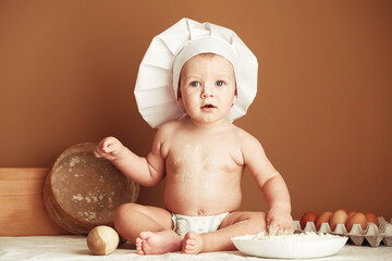 Little boy baker in a chef's hat sitting on the table playing with flour on a brown background with a wooden rolling pin, a round rustic sieve and eggs. Copy, empty space for text