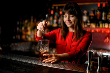 view on transparent glass with drink standing on bar and woman bartender is stirring it with spoon