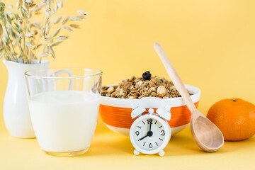 Healthy hearty breakfast. Baked granola in bowl, glass of milk, orange and alarm clock on yellow...