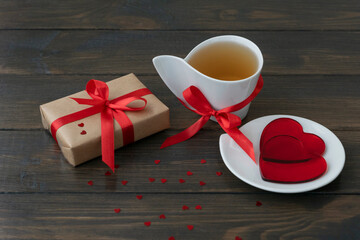 wrapped giftbox and white cup of tea tired up with red ribbons, two hearts lie on little plate. Valentines day concept