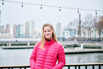 A pretty teenager is wearing a pink puffy coat and looks cosy and warm. She looks happy, has long fair hair and white skin with rosy cheeks and green eyes. The New York Skyline is in the background.