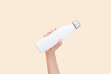 Close-up of hand holding white reusable steel thermo water bottle isolated on background of set sail champagne color, 2021 trend; with copy space.
