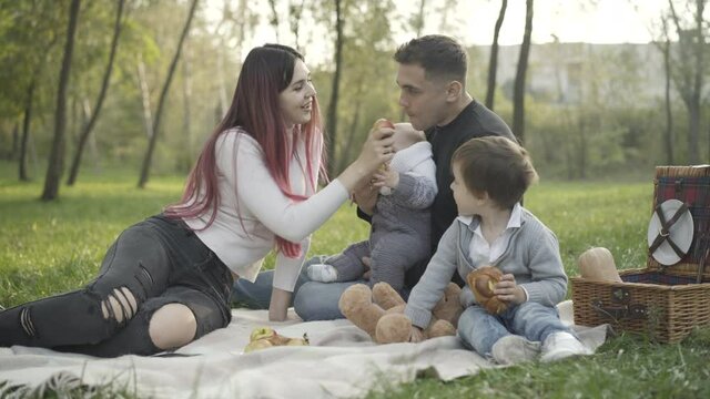 Carefree young Caucasian family sitting on blanket on summer meadow eating croissants and apples. Positive husband and wife enjoying picnic with little son and infant outdoors.