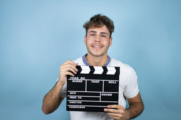 Young handsome man wearing a white t-shirt over blue background holding clapperboard very happy having fun