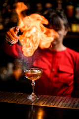 wine glass with iced drink stands on bar and lady barman sprinkles and sets fire over it