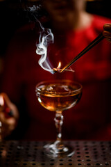 Close-up view of glass with drink over which bartender holds smoldering plant