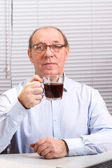 A man in the office with glasses at the computer drinks coffee.