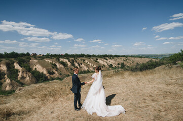 Fototapeta na wymiar Stylish groom in a blue suit and a beautiful curly-haired bride in a white dress stand in nature, against the background of hills and the sky, holding hands. Wedding portrait of newlyweds in love.