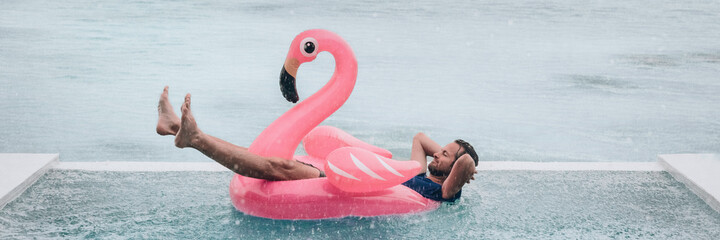 Happy man relaxing in swimming pool flamingo float despite bad rain weather. Travel summer vacation...