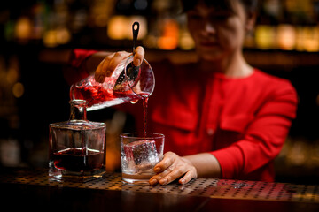 glass with large piece of ice stands on bar and bartender pours drink into it