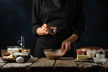 Pastry chef pours spaces into glass bowl for preparing dough on wooden table with variety of ingredients on dark blue background. Backstage of preparing sweet waffles. Frozen motion. Cooking process.