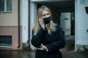 Fototapeta premium blonde business lady in black coat and mask on her face using smartphone against backdrop of arch of building. Lifestyle photo