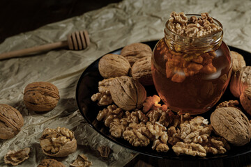 Honey and walnuts in a glass jar on a black plate. Background of crumpled paper and honey sticks.