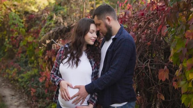 Pregnant couple shows a heart sign on the girl's belly standing in the autumn forest.