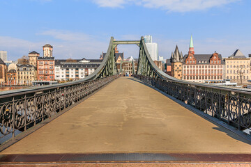 Obraz premium Historic footbridge over the Main in Frankfurt. Houses of the old town in the background with sunshine and a few clouds. Bridge with iron terrain