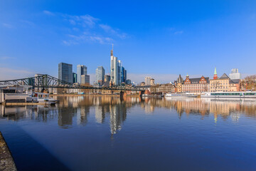 Fototapeta na wymiar Financial district of Frankfurt in sunshine. Skyline commercial buildings and bridge over the river Main with reflections. Ships at the moorings and historical buildings under blue sky