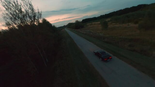 Black jdm car driving on countryside road on evening sunset background. Fpv drone cinematic view sport vehicle driving in twilight on suburban highway on summer trees and horizon landscape.