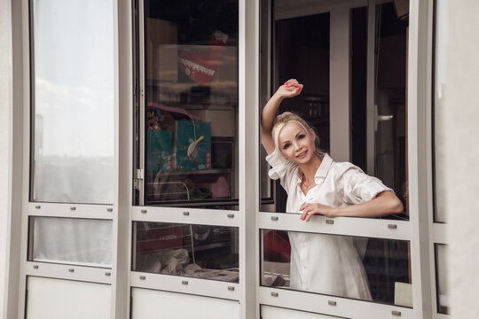Young woman in white men shirt looks out window and is emotional. Portrait of nice female. Slavic female in morning. Emotions and relax. Concept of homestay experiences and home comfort and waiting