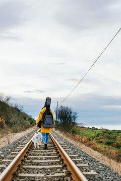 unrecognizable musician in a yellow coat walking her dog along the train tracks with the guitar loaded on his back. youth concept