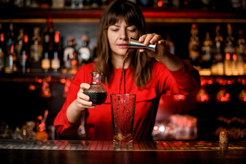 glass stands on bar and woman bartender accurate pours drink from jigger into it