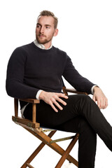 Attractive blonde man sitting down in front of a white background. Wearing dark pants and a dark sweater.
