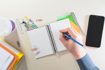 Top view of a male hand writing in a notebook, on a office desk with coffee cup, folders, supplies office and smart phone.