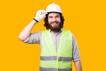 A nice portrait of a young bearded architect smiling at the camera is holding his white helmet near a yellow wall .