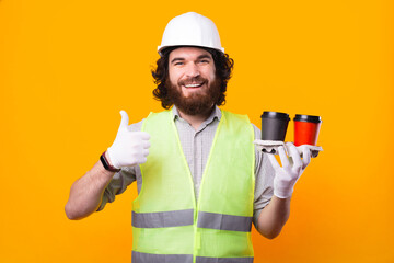 A young cheerful bearded engineer is looking at the camera smiling and holding two cups of hot drink is showing a thumb up that he likes the coffee .