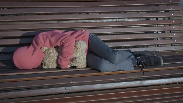Orphaned girl. An orphaned teen embrace her teddy bear and lay on the bench.
