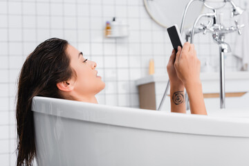 young tattooed woman using smartphone while taking bath