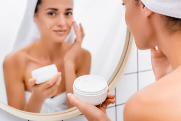 Obraz na płótnie Canvas young woman applying face cream near blurred mirror reflection in bathroom