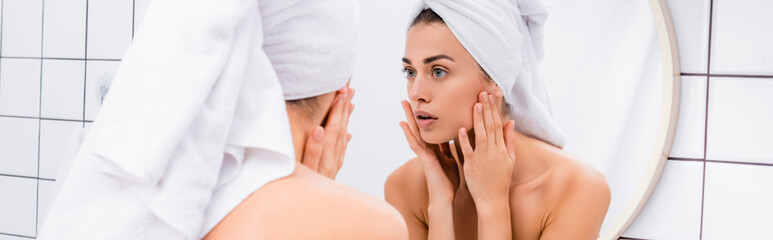 displeased young woman with towel on head looking in mirror and touching face in bathroom, banner