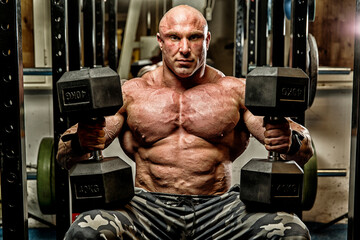 Strong man with huge dumbbells in the gym shirtless with muscular torso.