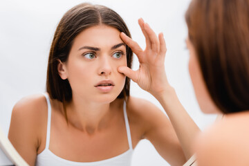 worried young woman looking in mirror and touching face, blurred foreground