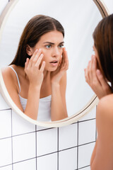 worried young woman looking in mirror and touching face in bathroom, blurred foreground