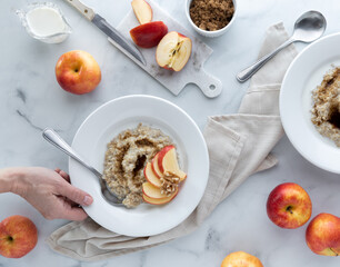 Hand holding a bowl of steel cut oats with brown sugar and apple slices against a light background.