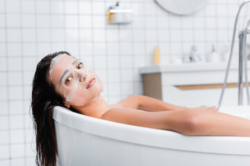 young woman in face mask relaxing in bathtub and looking at camera