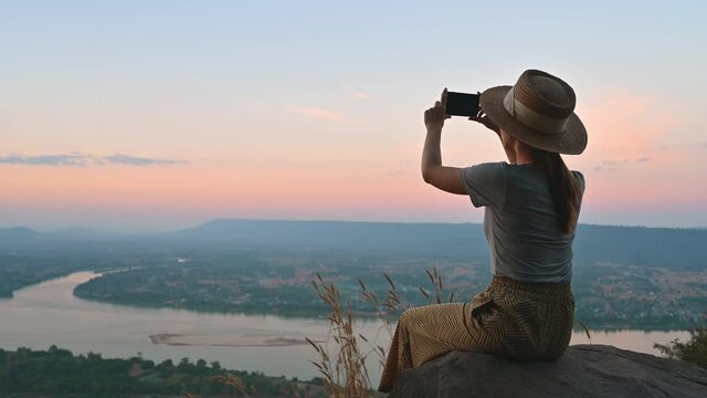 Asian woman sitting on the cliff of Pha tak suea temple photographing the view of Mekong river between Thailand and Laos in evening