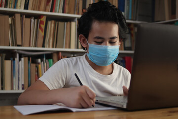 Asian boy student wearing protective mask learning studying online with laptop computer in library, school from home