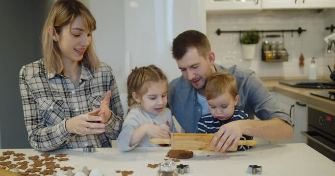 Young family spending time at home together. Parents have fun with children baking christmas cookies pastry in the kitchen. Mother and father teach their daughter and son to to roll out the dough.