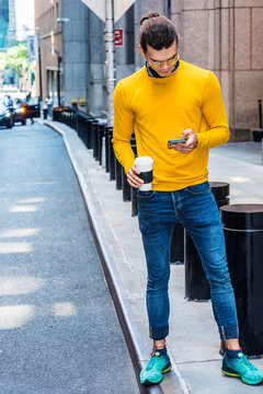 Young Hispanic American with hair bun, wearing glasses, yellow long sleeve T shirt, blue jeans, green sneakers, holding cup of coffee, standing on street in New York City, texting on cell phone..