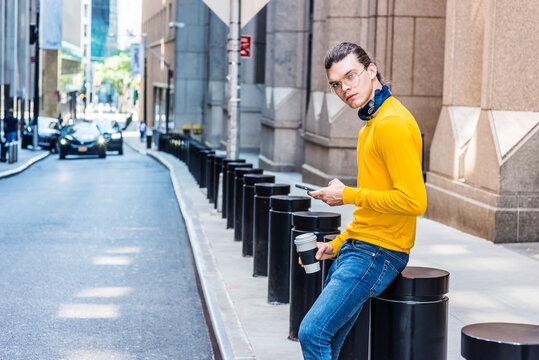 Young Hispanic American with hair bun, wearing glasses, yellow long sleeve T shirt, blue jeans, small black scarf around neck, holding cup of coffee, sitting on street in New York City, texting..