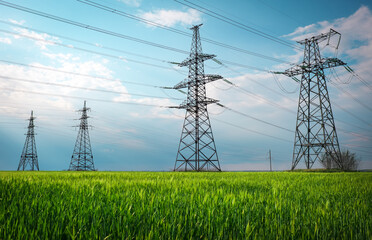 High voltage lines and power pylons in a flat and green agricultural landscape on a sunny day with clouds in the blue sky. Cloudy and rainy. Wheat is growing