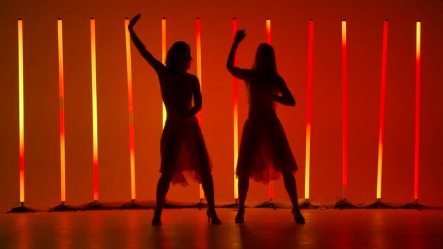 Two graceful young women practice the elements of salsa dance in a dark studio against a background of bright red orange neon lights. Silhouettes. Slow motion.